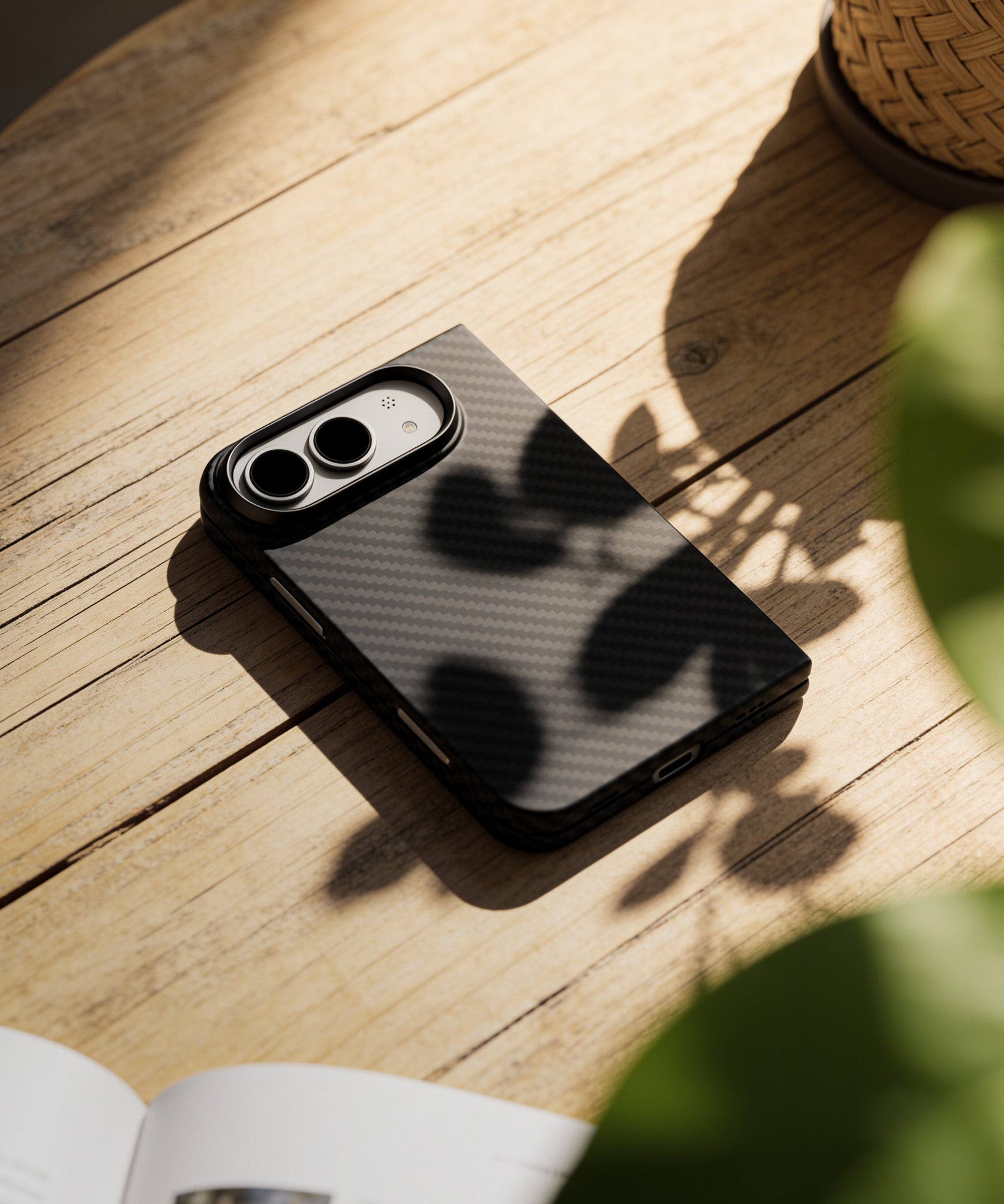 A black carbon-fiber-textured phone case with a camera bar cutout, resting on a rustic wooden table with natural shadows from nearby leaves.