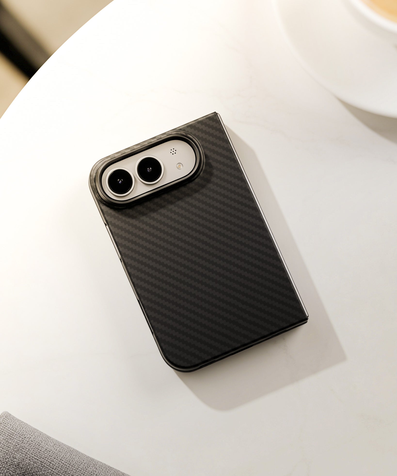 A black carbon fiber textured phone case with a silver camera bumper, shown on a white marble tabletop with a coffee cup in the background.
